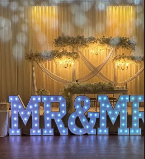 Lettres lumineuses MR & MRS devant une draperie dorée et des guirlandes florales