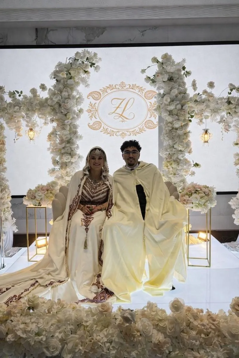 Mariés en tenues traditionnelles devant un backdrop floral avec monogramme