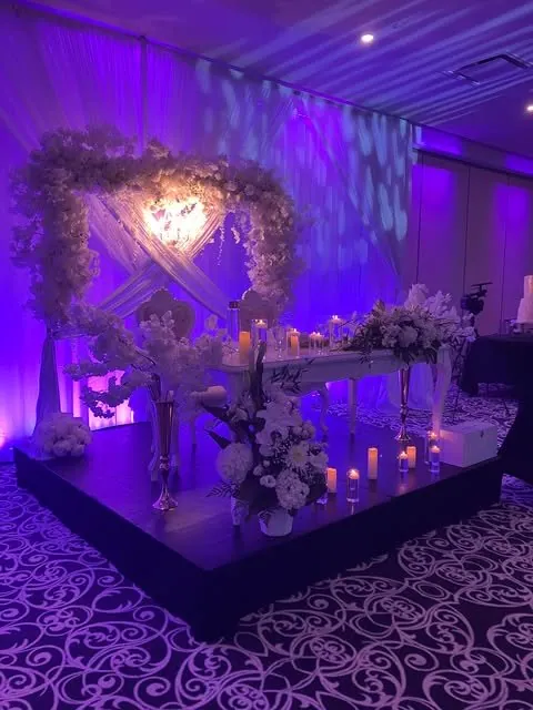 Table d’honneur blanche, drapé croisé et cadre floral sous éclairage mauve